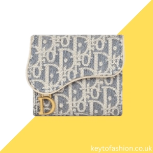 Dior Wallet Worth the Splurge? Exploring Luxury, Investment, and Practicality 2 A grey and white Dior monogram wallet, likely the Dior Saddle Lotus Wallet, featuring the iconic "CD" logo pattern. The wallet has a curved flap closure with a gold-toned 'D' charm hanging from it. The background is split diagonally, with the top left being light blue and the bottom right being yellow.