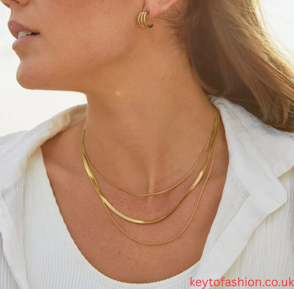 Layered Chains Your 2025 Style Obsession 3 A close-up shot of a person wearing a white collared shirt and three delicate gold chain necklaces of varying lengths. The chains are a snake chain style. The person also wears a small gold hoop earring and has long, light brown hair. The image is bright and airy, with a focus on the jewelry and the person's neck and shoulder area.
