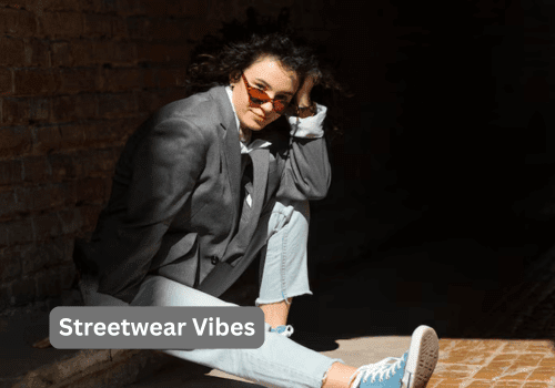How to Style a Silver Outfit in 2025 and Shine with Confidence 4 A person with curly dark hair wearing sunglasses, a gray oversized blazer, a white hoodie, light blue jeans, and blue sneakers sits on the ground against a brick wall, with a dark shadow obscuring the right side of the image. A gray box with the text "Streetwear Vibes" is in the bottom left corner.