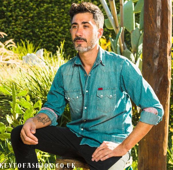 How to Style Linen Shirts for Men in 2025 3 A stylish man with a salt-and-pepper beard and mustache, wearing a distressed denim shirt and black pants, is sitting outdoors. He has a tattoo on his left forearm and is looking slightly to the left of the camera with a serious expression. The background is a garden with various plants, including a cactus and a wooden post. There is a watermark in the bottom left corner that reads "KEYTOFASHION.CO.UK".