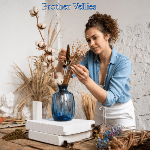 10 Remarkable Brands Like Idioma Every Style Enthusiast Will Fall in Love With in 2025 8 A woman with curly brown hair wearing a light denim shirt and white pants is arranging dried flowers and cotton stems in a blue glass vase. She is standing in a brightly lit room with white brick walls.