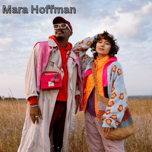 10 Remarkable Brands Like Idioma Every Style Enthusiast Will Fall in Love With in 2025 9 A man and a woman are standing together in a field of tall grass. The man, on the left, is wearing a long light-colored trench coat, a red hoodie, a red cap, and sunglasses. The woman, on the right, has short curly hair and is wearing a light blue and pink floral cardigan over an orange vest and a purple top. She is carrying a large striped woven bag.