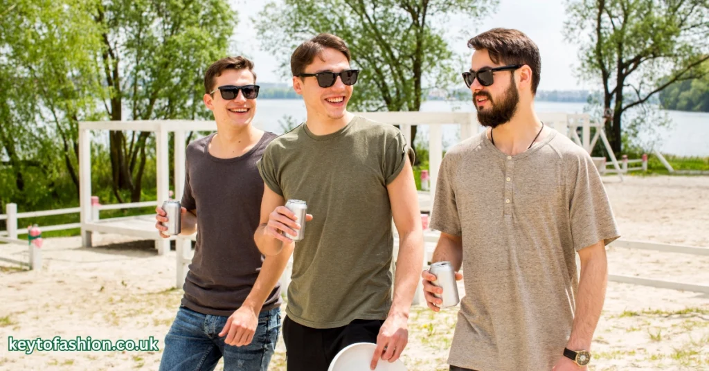 Three young men are walking outdoors in a park-like setting on a sunny day. The man on the left is wearing a dark grey t-shirt and sunglasses, holding a silver can. The man in the middle, also wearing sunglasses, is smiling and holding a silver can, wearing an olive green t-shirt. The man on the right, with a beard and sunglasses, is wearing a light brown or tan henley shirt and holding a silver can. In the background, there are trees and a body of water.