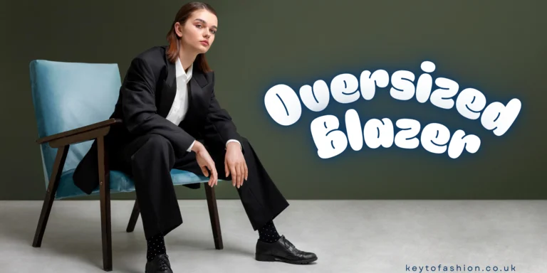 How to Wear an Oversized Blazer in 2025 and Feel Like a Total Boss 9 A woman with a brown bob hairstyle is seated on a light blue armchair against a dark green background. She is wearing an oversized black blazer, black wide-leg trousers, a white shirt, black shoes, and black socks with white polka dots. The words "Oversized Blazer" are written in a large, white, puffy font with a blue outline to the right of her. The website "keytofashion.co.uk" is written in small white text at the bottom right corner.