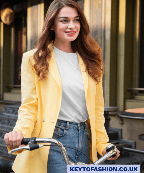 How to Wear an Oversized Blazer in 2025 and Feel Like a Total Boss 3 A young woman with long, wavy brown hair is smiling and looking to her right while holding the handlebars of a bicycle. She is wearing a pale yellow oversized blazer over a simple white t-shirt and light-wash blue jeans. The background is a blurred outdoor scene with buildings and stairs. The overall tone is cheerful and stylish.