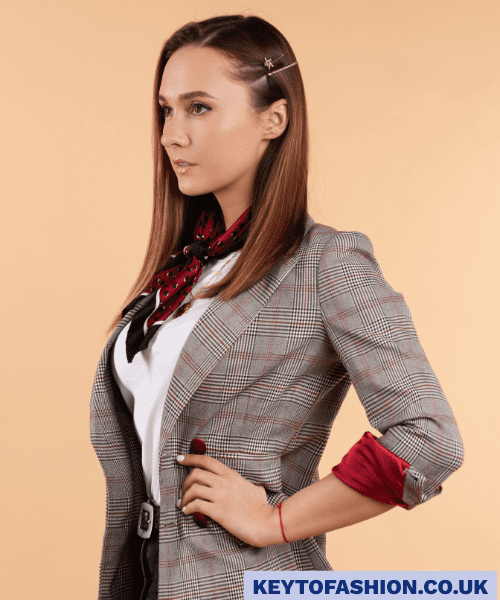 How to Wear an Oversized Blazer in 2025 and Feel Like a Total Boss 4 A young woman with straight brown hair, styled with a star-shaped hair clip, is posing against a plain beige background. She is wearing a grey and red plaid blazer with the sleeves cuffed to reveal a solid red lining. Underneath, she has on a white t-shirt and a small red and black patterned scarf tied around her neck. She is looking to her left with a serious expression, and her left hand is on her hip.