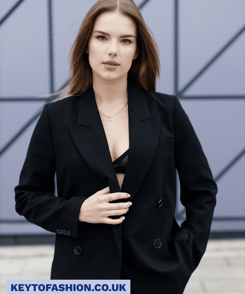 How to Wear an Oversized Blazer in 2025 and Feel Like a Total Boss 5 A stylish young woman with long brown hair looks directly at the camera. She is wearing a black double-breasted blazer, unbuttoned to reveal a glimpse of a black lace bra and a delicate gold necklace. Her left hand is resting on the blazer's lapel. The background is a modern, geometric wall in shades of grey and purple. The overall mood is confident and sophisticated.