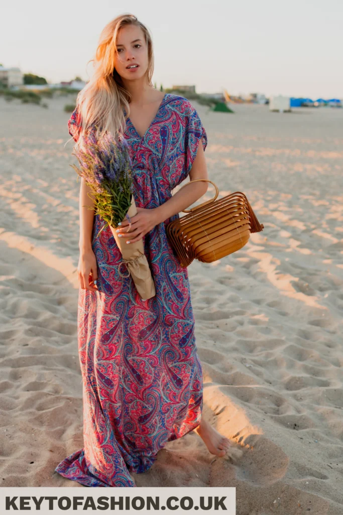 How to Master Styling a Boho Maxi Dress Floral Look The Secret to Natural Polished Dressing 4 A blonde woman in a flowing, paisley-print maxi dress stands barefoot on a sandy beach at sunset, holding a bouquet of lavender and a wood-slat tote bag.