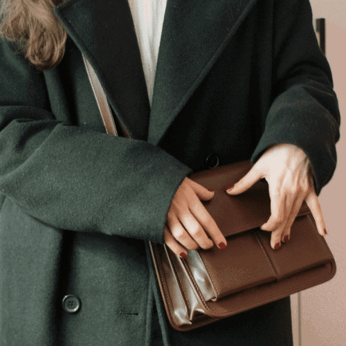 The Art of Accessory Design and How Small Details Define Big Fashion Statements 3 A close-up of a person wearing a black coat and holding a brown leather satchel bag with both hands.