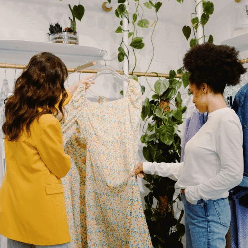 The Art of Accessory Design and How Small Details Define Big Fashion Statements 4 Two women are shopping in a boutique. One woman in a yellow blazer holds up a floral dress on a hanger for the other woman to see.